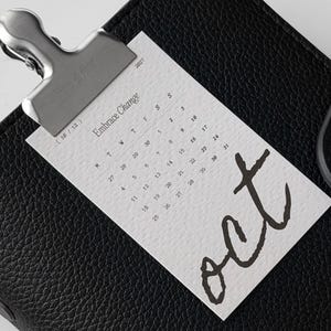 May include: A black textured leather planner secured with a silver metal clip holding a white calendar card for October 2027. The card features the text Embrace Change, the month abbreviation oct in large script, and a grid of dates for the month.
