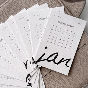 May include: A set of off-white paper calendar cards arranged in a fan shape, each featuring the text Begin with Intention, 2027, and a monthly grid. The cards display bold black handwritten-style lettering at the bottom and are placed on a textured taupe surface.