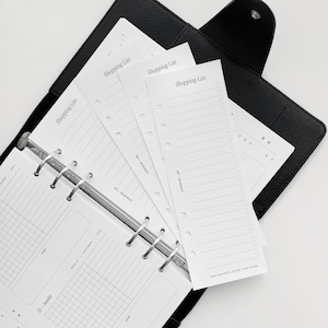 May include: A black leather bound planner with a silver ring binder. The planner is open to a weekly spread with a grid layout. There are several white shopping list inserts with the text "Shopping List" printed on them.