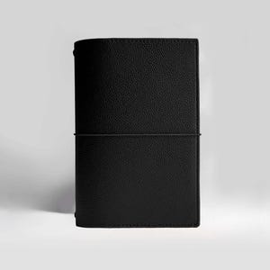 May include: A black leather journal with a textured surface. The journal is closed with an elastic band. The cover is a solid black color. The journal is standing upright against a white background.