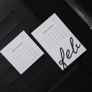 May include: Two white rectangular calendar cards placed inside a black textured leather binder. The left card is labeled 1/12 Begin with Intention, and the right card is labeled 2/12 Choose Connection with the handwritten word Feb in black ink.