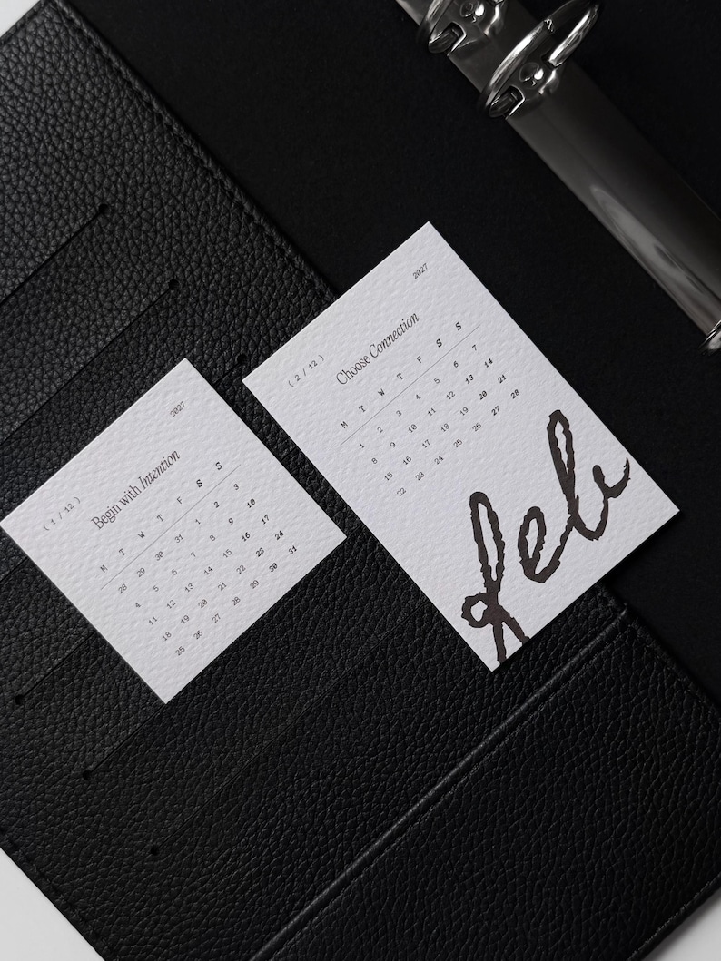 May include: Two white rectangular calendar cards placed inside a black textured leather binder. The left card is labeled 1/12 Begin with Intention, and the right card is labeled 2/12 Choose Connection with the handwritten word Feb in black ink.