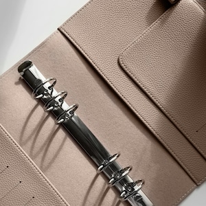 May include: A light beige leather binder with a silver metal ring mechanism. The binder has a pocket on the front cover and a flap on the back cover.