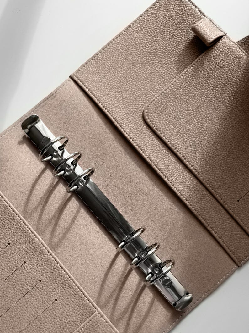 May include: A light beige leather binder with a silver metal ring mechanism. The binder has a pocket on the front cover and a flap on the back cover.