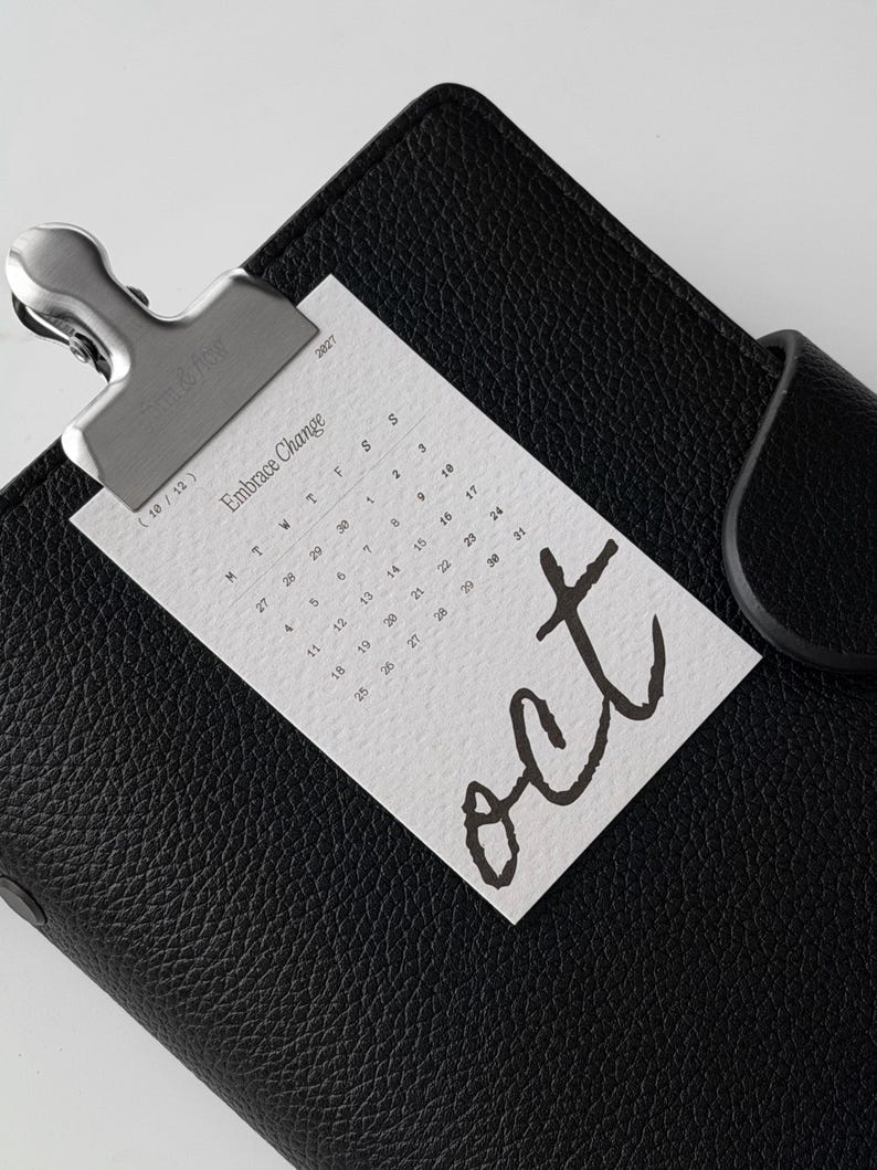 May include: A black textured leather planner secured with a silver metal clip holding a white calendar card for October 2027. The card features the text Embrace Change, the month abbreviation oct in large script, and a grid of dates for the month.