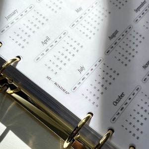 Pu&ograve; includere: Un inserto per agenda in carta bianca con un calendario 2024 che mostra i mesi da gennaio a novembre in testo nero. Le pagine sono tenute insieme da un meccanismo di rilegatura a sei anelli in metallo color oro. Testo MY ROADMAP visibile.
