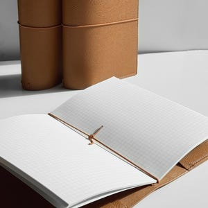 K&ouml;nnte beinhalten: Offenes, hellbraunes Lederjournal mit karierten Seiten. Das Journal wird mit einem braunen Gummiband verschlossen. Zwei weitere Journale sind im Hintergrund zu sehen. Das Leder hat eine strukturierte Oberfl&auml;che.