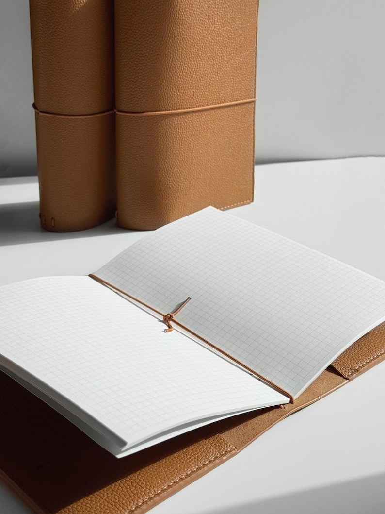 K&ouml;nnte beinhalten: Offenes, hellbraunes Lederjournal mit karierten Seiten. Das Journal wird mit einem braunen Gummiband verschlossen. Zwei weitere Journale sind im Hintergrund zu sehen. Das Leder hat eine strukturierte Oberfl&auml;che.