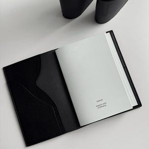 May include: Open black leather notebook with a textured cover and a pocket on the left side. The notebook is open to a blank page with text in the upper right corner. A pair of black shoes are in the background.