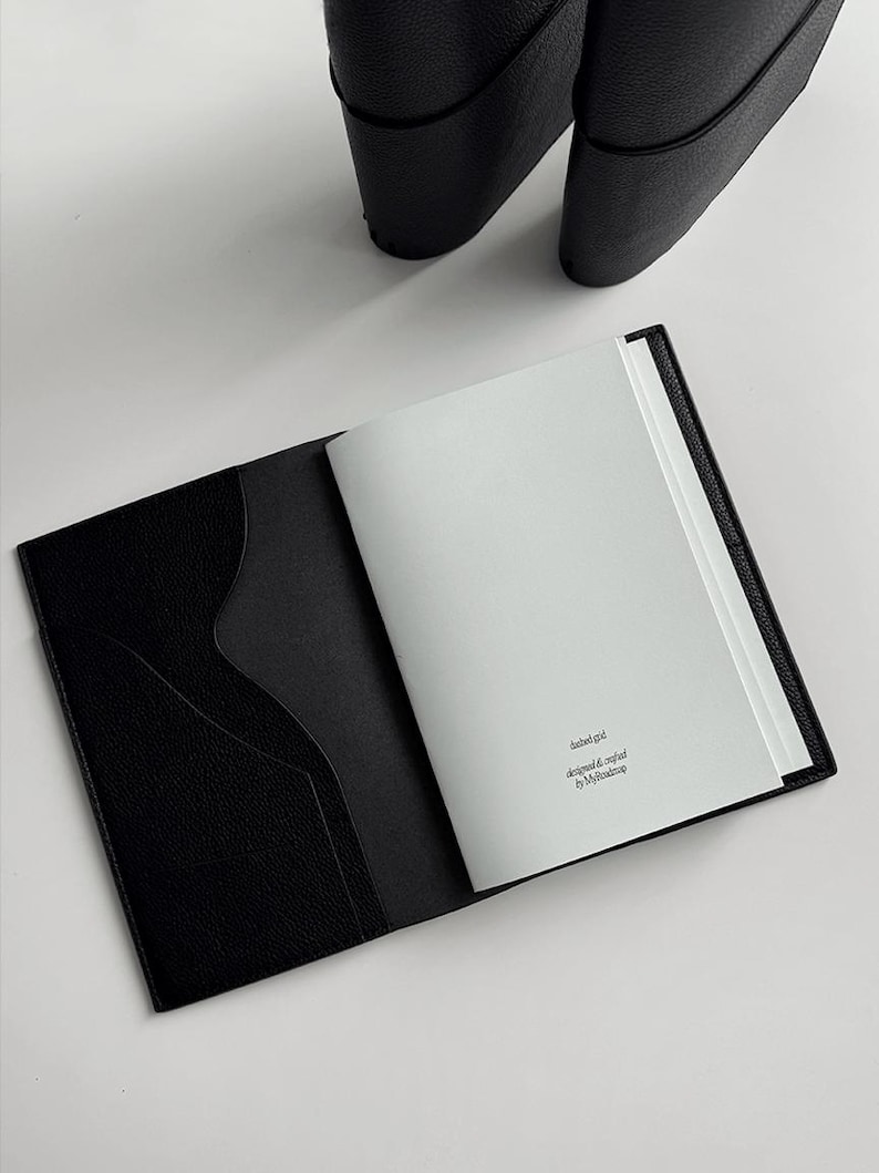 May include: Open black leather notebook with a textured cover and a pocket on the left side. The notebook is open to a blank page with text in the upper right corner. A pair of black shoes are in the background.