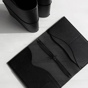 May include: An open black leather journal with a textured cover and interior pockets. The journal is held open by a black elastic cord. Two black leather cup holders are in the background.