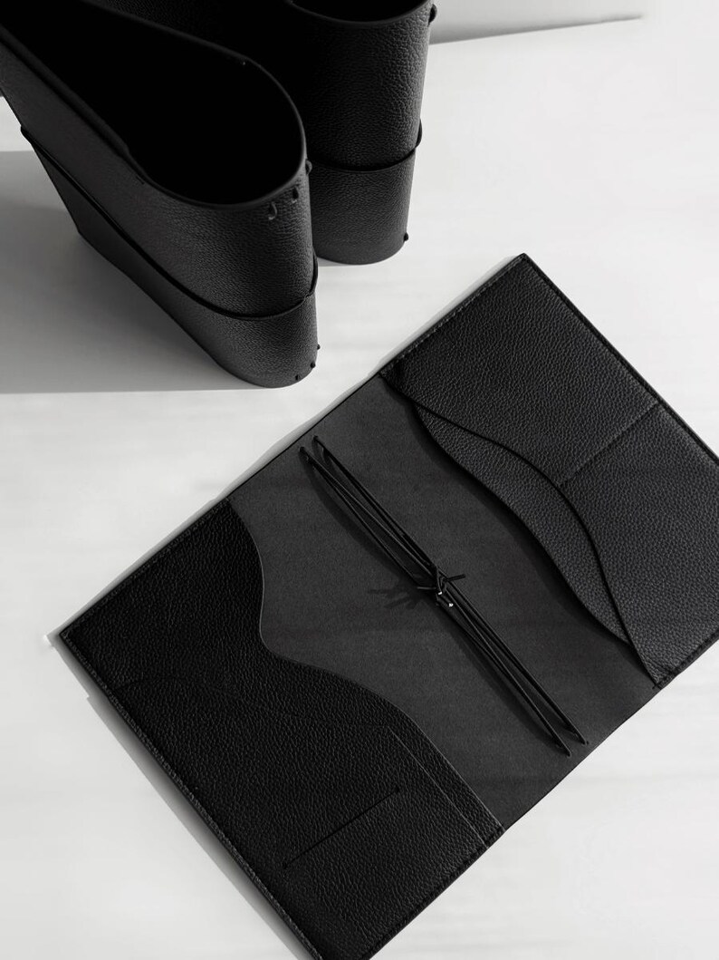 May include: An open black leather journal with a textured cover and interior pockets. The journal is held open by a black elastic cord. Two black leather cup holders are in the background.