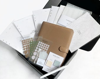 May include: A black box filled with planner accessories, including a brown leather planner, white paper, stickers, and a pen. The box is open and the items are visible inside.