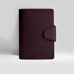 May include: A dark burgundy leather journal with a textured finish and a curved flap closure. The journal is closed and features the embossed text "MY ROADMAP" at the bottom. It is set against a plain white backdrop.