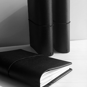 May include: Black leather journals with a textured finish. One journal is open, revealing white pages, while two stand upright. A black elastic band secures each journal. The journals are on a white surface.