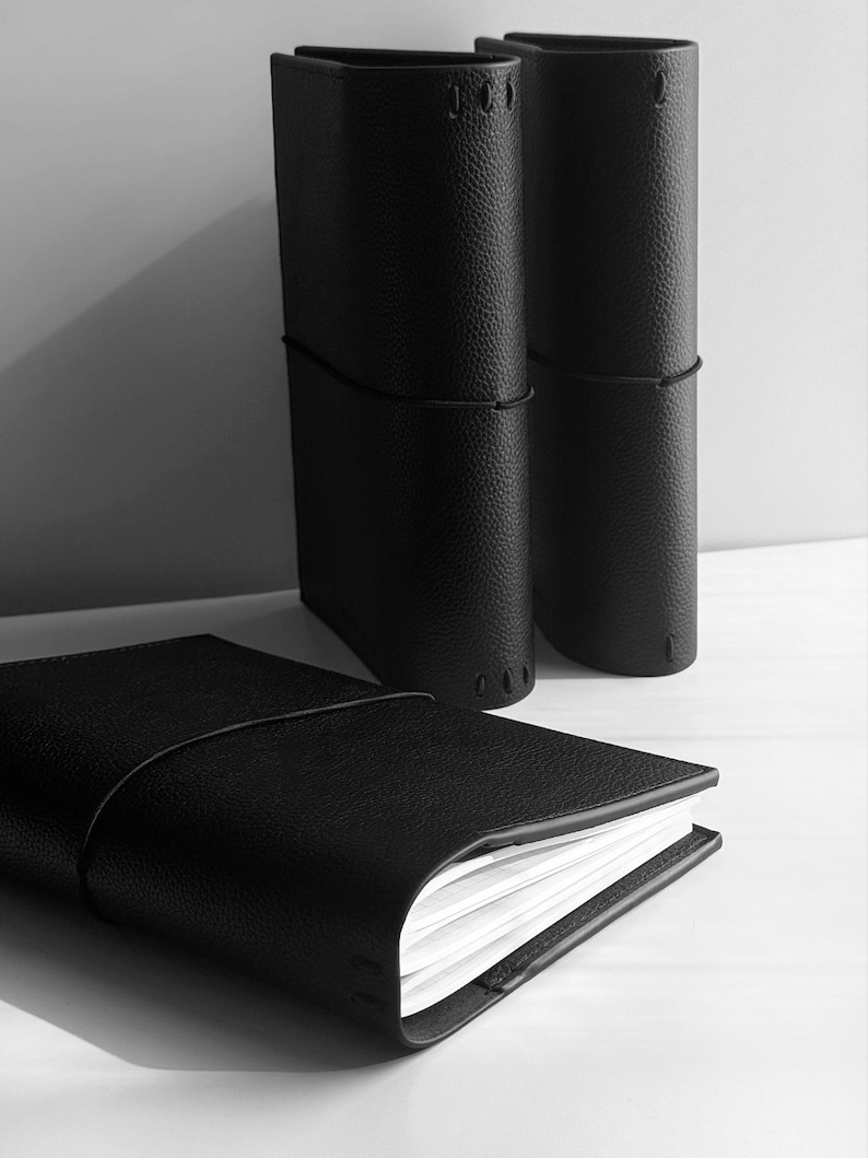 May include: Black leather journals with a textured finish. One journal is open, revealing white pages, while two stand upright. A black elastic band secures each journal. The journals are on a white surface.