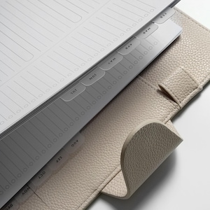 May include: A beige textured leather planner with a metal ring binder mechanism. The interior features white paper pages with lined sections and monthly tab dividers labeled from February through December in black sans-serif capital letters.