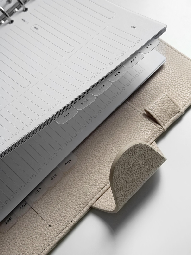 May include: A beige textured leather planner with a metal ring binder mechanism. The interior features white paper pages with lined sections and monthly tab dividers labeled from February through December in black sans-serif capital letters.