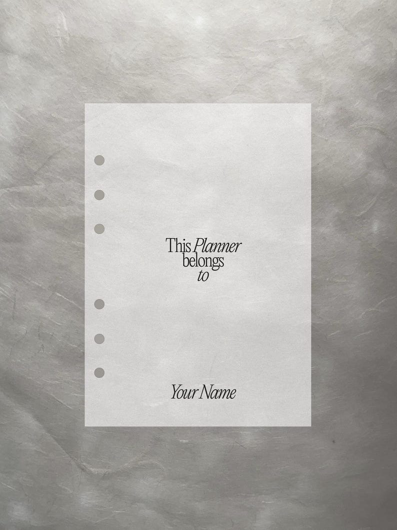 May include: A white planner page with pre-punched holes. The text "This Planner belongs to Your Name" is printed in a classic serif font. The page is set against a textured, silver-gray background.