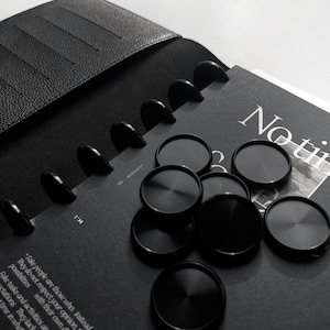 Könnte beinhalten: Ein schwarzes, ledergebundenes Notizbuch mit schwarzem Einband und schwarzem Gummibandverschluss. Das Notizbuch hat eine schwarz-weiße Seite mit dem aufgedruckten Text "No time". Das Notizbuch ist geöffnet und mehrere schwarze Metall-Disc-Ringe halten die Seiten zusammen. Mehrere schwarze Metall-Discs liegen verstreut auf der Seite.