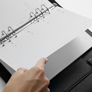 May include: A black leather-textured binder containing white planner pages with printed sections for daily scheduling, a roadmap, and lined note areas. The binder features a silver-colored metal ring mechanism that holds the paper inserts securely in place.