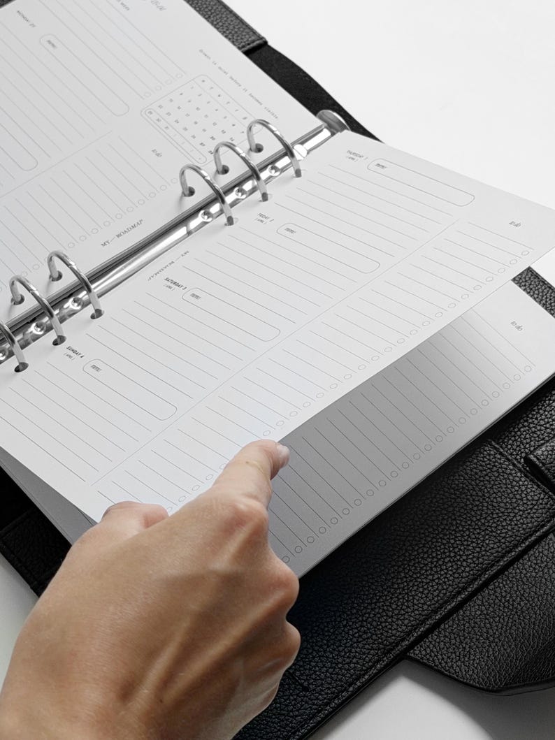 May include: A black leather-textured binder containing white planner pages with printed sections for daily scheduling, a roadmap, and lined note areas. The binder features a silver-colored metal ring mechanism that holds the paper inserts securely in place.