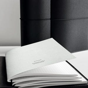May include: An open black leather journal with a grid-lined paper insert. The pages are white with a subtle grid pattern. The text "dashed grid designed & crafted by MyRoadmap" is printed on the top page. A second closed journal is in the background.