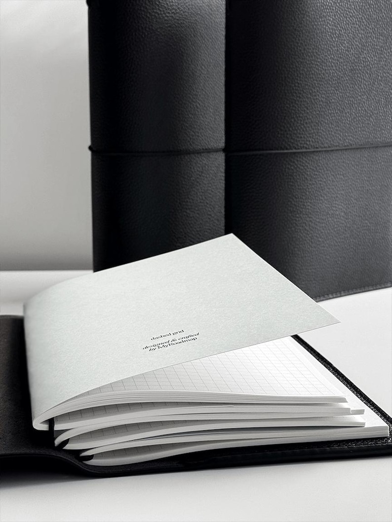 May include: An open black leather journal with a grid-lined paper insert. The pages are white with a subtle grid pattern. The text "dashed grid designed & crafted by MyRoadmap" is printed on the top page. A second closed journal is in the background.