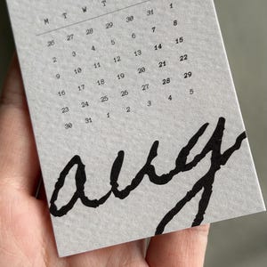 May include: A light gray paper calendar page for August, labeled Let Go and (8/12), featuring a grid of dates from Monday to Sunday. The word aug is printed in large, bold, black, handwritten-style script across the bottom portion of the page.
