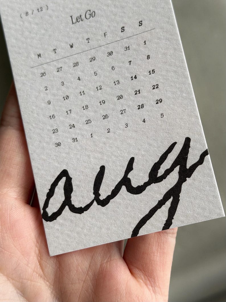 May include: A light gray paper calendar page for August, labeled Let Go and (8/12), featuring a grid of dates from Monday to Sunday. The word aug is printed in large, bold, black, handwritten-style script across the bottom portion of the page.