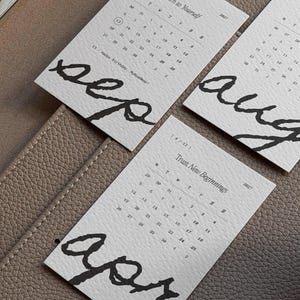 May include: Three rectangular white paper calendar cards for 2027. Each card features a month name in large black handwritten script, a grid of dates, and a small phrase like Return to Yourself or Trust New Beginnings printed in black ink.