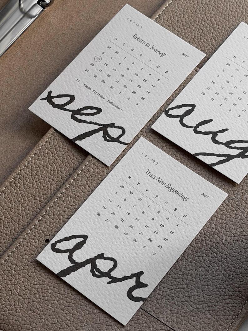 May include: Three rectangular white paper calendar cards for 2027. Each card features a month name in large black handwritten script, a grid of dates, and a small phrase like Return to Yourself or Trust New Beginnings printed in black ink.