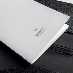 K&ouml;nnte beinhalten: Ein wei&szlig;es, offenes Notizbuch mit dem Text "dotted designed & crafted by MyRoadmap" in einem schwarzen Ledereinband. Das Notizbuch wird mit einem schwarzen Gummiband offen gehalten.