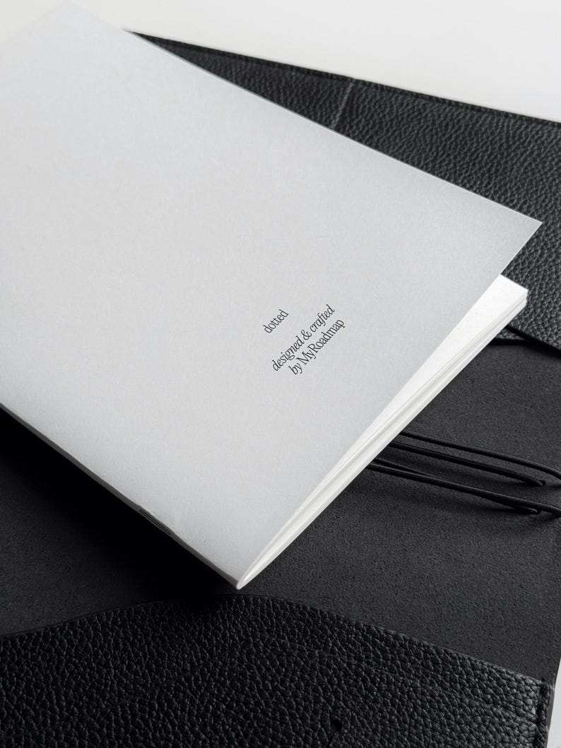K&ouml;nnte beinhalten: Ein wei&szlig;es, offenes Notizbuch mit dem Text "dotted designed & crafted by MyRoadmap" in einem schwarzen Ledereinband. Das Notizbuch wird mit einem schwarzen Gummiband offen gehalten.