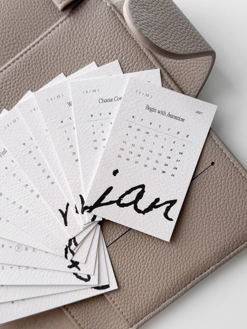 May include: A set of off-white paper calendar cards arranged in a fan shape, each featuring the text Begin with Intention, 2027, and a monthly grid. The cards display bold black handwritten-style lettering at the bottom and are placed on a textured taupe surface.