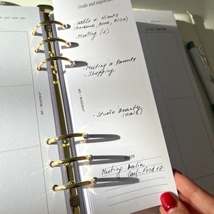 Könnte beinhalten: Ein weißer Planer mit goldenen Ringen. Der Planer hat eine Seite geöffnet, die mit "Ziele und wichtig" beschriftet ist. Die Seite listet mehrere Aufgaben auf, darunter "Anrufe mit Kunden (Barbara, Anna, Alisa)", "Meeting (2)", "Treffen mit den Eltern", "Einkaufen", "Studio Beauty" und "Notizen". Der Notizbereich enthält den Text "Meeting: Berlin, Cal-Nord 17".