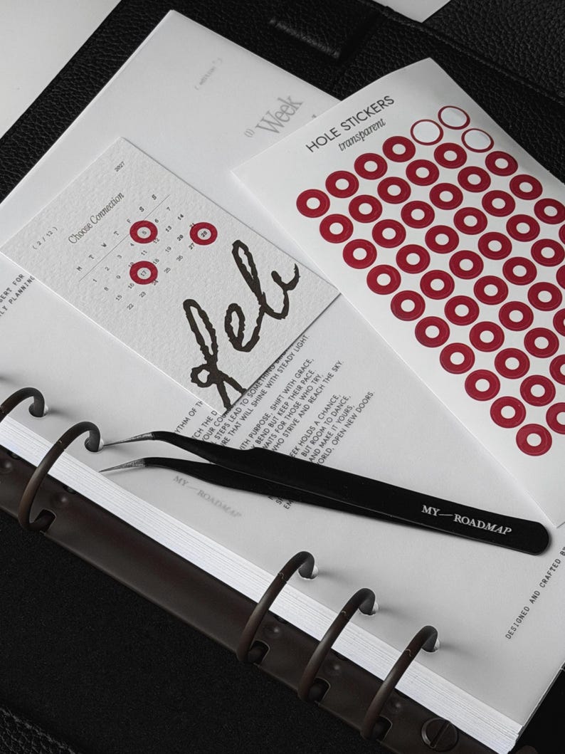 May include: A black metal pair of tweezers with the text MY ROADMAP printed on the handle, resting on a white planner page. Beside it is a sheet of transparent red ring-shaped hole stickers and a calendar card labeled Feb with red circles.