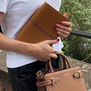 K&ouml;nnte beinhalten: Ein braunes Leder-Portfolio mit blauem Gummiband und eine braune Lederhandtasche mit goldfarbener Hardware. Die Handtasche hat einen oberen Griff und einen Schulterriemen. Das Portfolio und die Handtasche sind aus strukturiertem Leder.