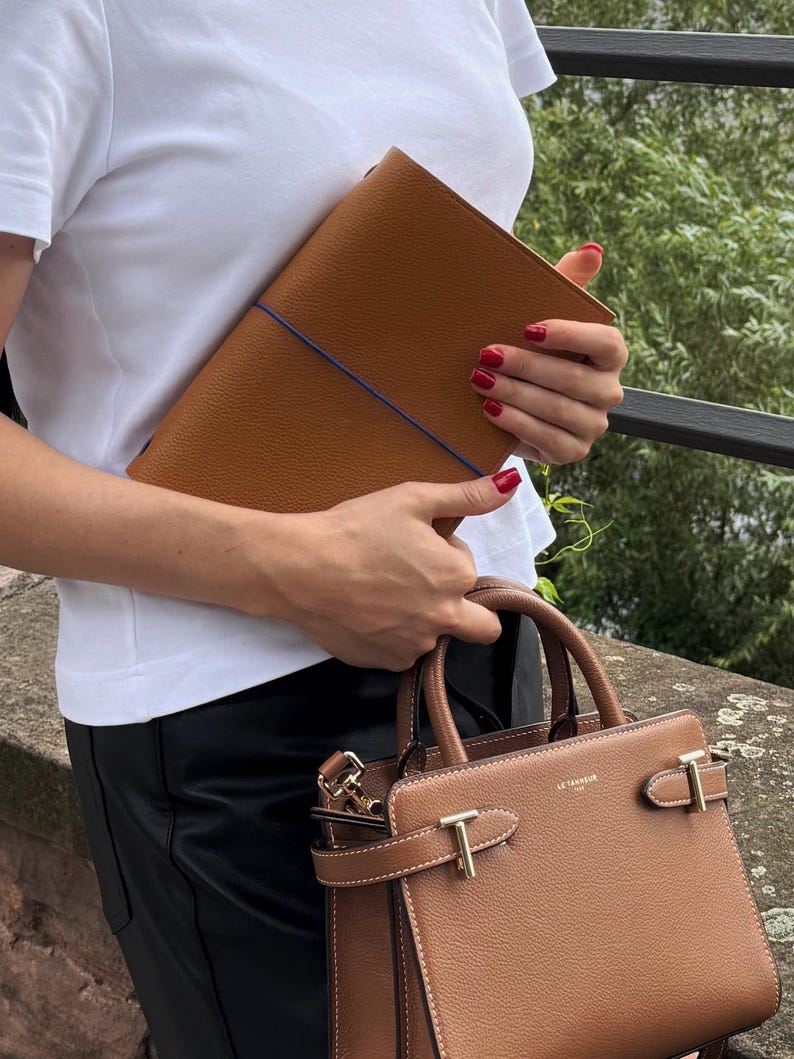 K&ouml;nnte beinhalten: Ein braunes Leder-Portfolio mit blauem Gummiband und eine braune Lederhandtasche mit goldfarbener Hardware. Die Handtasche hat einen oberen Griff und einen Schulterriemen. Das Portfolio und die Handtasche sind aus strukturiertem Leder.