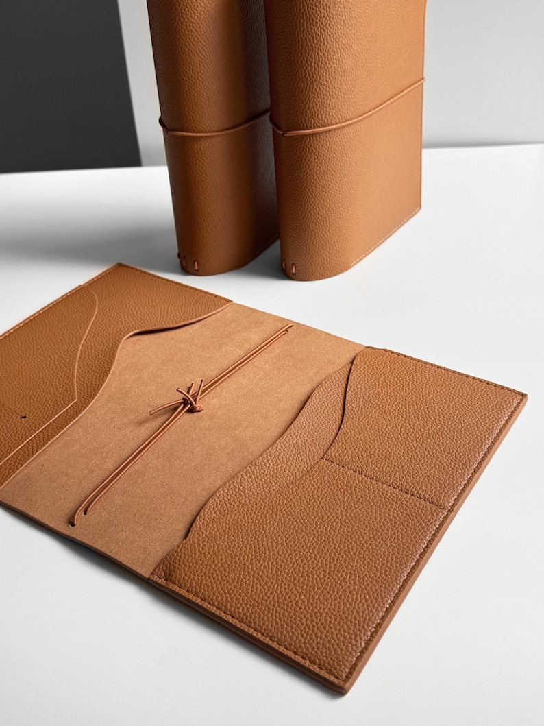 K&ouml;nnte beinhalten: Offenes hellbraunes Lederjournal mit Taschen und Kordelverschluss. Zwei weitere Journale stehen im Hintergrund aufrecht. Die Journale haben eine strukturierte Oberfl&auml;che und eine neutrale Farbpalette.