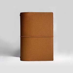 K&ouml;nnte beinhalten: Ein braunes Lederjournal mit strukturierter Oberfl&auml;che. Das Journal wird mit einem passenden Gummiband gesichert. Der Einband ist rechteckig mit abgerundeten Kanten. Das Journal ist etwa 20 cm hoch und 13 cm breit.