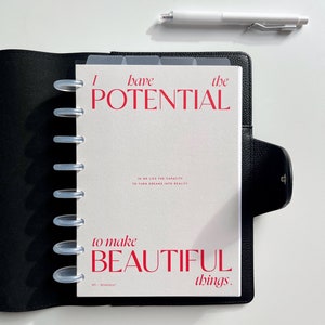 Może przedstawiać: Czarny sk&oacute;rzany planer z białym wkładem z napisem "I have the POTENTIAL to make BEAUTIFUL things." Biały długopis spoczywa na białej powierzchni. Planer ma przezroczyste przegrody i srebrne pierścienie.
