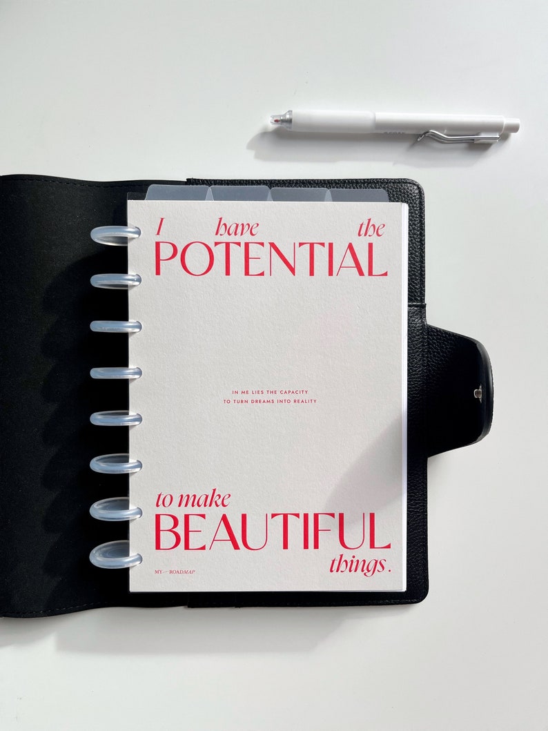 Może przedstawiać: Czarny sk&oacute;rzany planer z białym wkładem z napisem "I have the POTENTIAL to make BEAUTIFUL things." Biały długopis spoczywa na białej powierzchni. Planer ma przezroczyste przegrody i srebrne pierścienie.