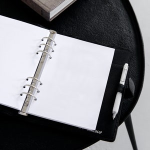 May include: An open black leather binder with blank white paper, a silver pen, and a gray book on a black table. The binder has a silver ring mechanism and a pen holder. The text "ROADMAP" is visible on the binder.