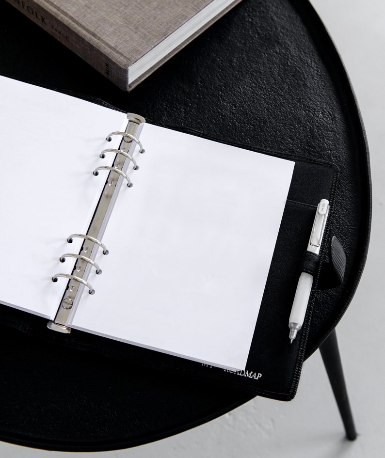 May include: An open black leather binder with blank white paper, a silver pen, and a gray book on a black table. The binder has a silver ring mechanism and a pen holder. The text "ROADMAP" is visible on the binder.