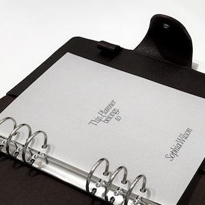 May include: A dark brown leather planner with silver metal rings. The planner is open, revealing a white sheet of paper with the text "This Planner belongs to" and the name "Sophia Wilson" printed on it.