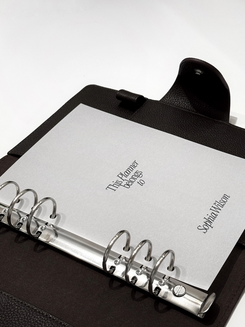 May include: A dark brown leather planner with silver metal rings. The planner is open, revealing a white sheet of paper with the text "This Planner belongs to" and the name "Sophia Wilson" printed on it.