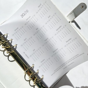 Pu&ograve; includere: Un'agenda ad anelli bianca con una pagina di calendario 2024 che mostra i mesi da gennaio a dicembre. La pagina include il testo MY ROADMAP e una citazione di Booker T. Washington sul successo. L'agenda ha anelli in metallo color oro.