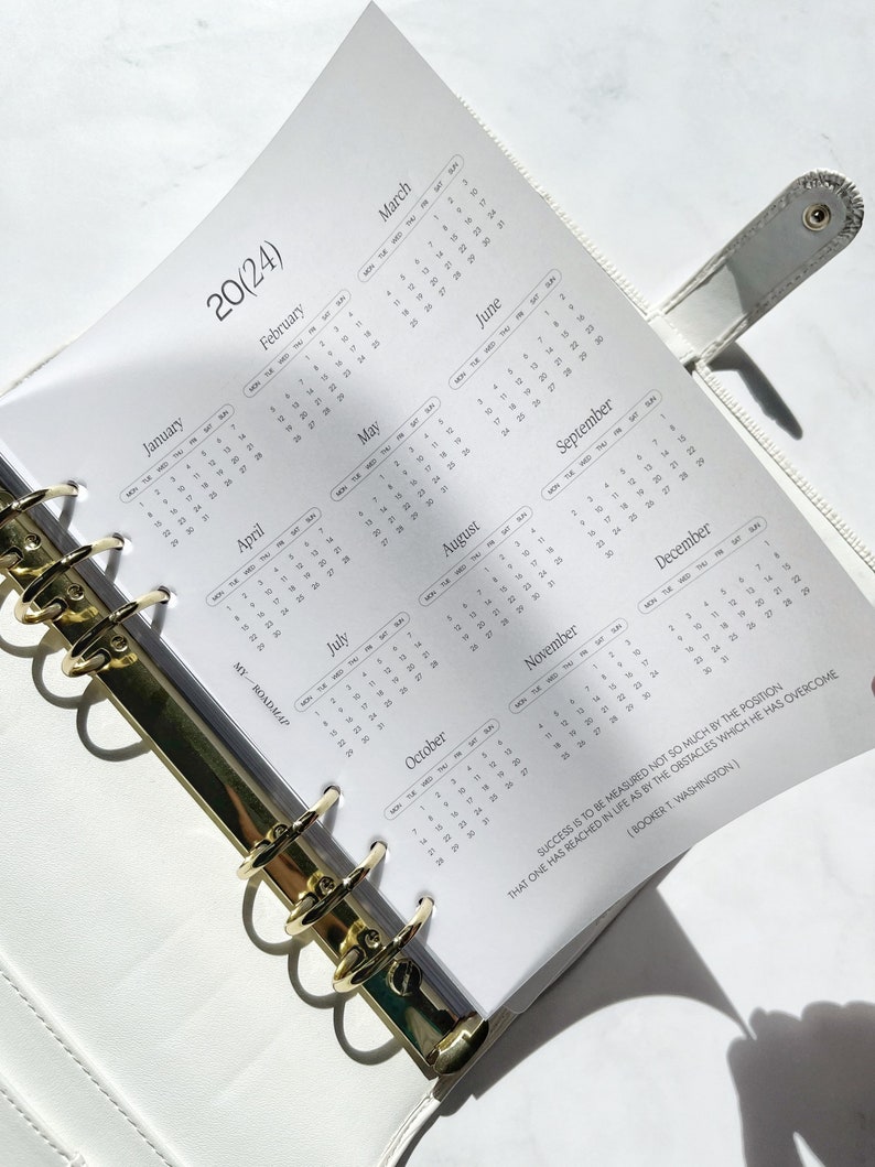 Pu&ograve; includere: Un'agenda ad anelli bianca con una pagina di calendario 2024 che mostra i mesi da gennaio a dicembre. La pagina include il testo MY ROADMAP e una citazione di Booker T. Washington sul successo. L'agenda ha anelli in metallo color oro.
