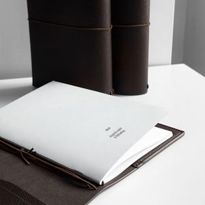 K&ouml;nnte beinhalten: Ein braunes Lederjournal mit passendem Lederbezug, ge&ouml;ffnet, um leere wei&szlig;e Seiten freizugeben. Zwei weitere Journale stehen im Hintergrund aufrecht. Der Text "brial designed & crafted by Mjolkman" ist auf der Seite aufgedruckt.